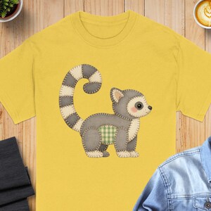 May include: Yellow t-shirt with a cartoon lemur illustration. The lemur is grey with a striped tail and a green plaid belly. The design has a stitched, handmade appearance.
