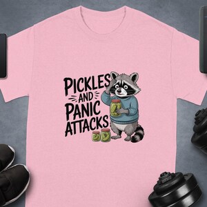 May include: Pale pink t-shirt featuring a cartoon raccoon clutching a jar of pickles, with the words "Pickles and Panic Attacks". The raccoon is wearing a blue sweater. Additional pickle jars are visible.