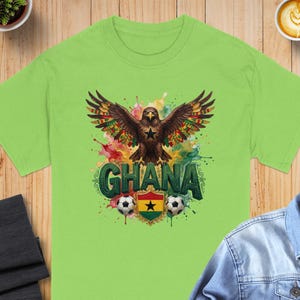 May include: A lime green t-shirt featuring a graphic design with a brown eagle with outstretched wings, the word "GHANA", soccer balls, and the Ghana flag. The design has colorful accents.