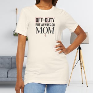 May include: Cream-colored t-shirt with the text "OFF-DUTY BUT ALWAYS ON MOM" in black and pink lettering. A lightning bolt graphic is below the word "MOM". The shirt is being worn by a person.