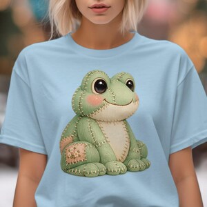 Könnte beinhalten: Hellblaues T-Shirt mit einem aufgenähten Frosch-Design. Der Frosch ist grün mit rosa Wangen, großen schwarzen Augen und einem lächelnden Gesichtsausdruck. Das Design hat eine Patchwork-Ästhetik mit sichtbaren Nähten und Patches.