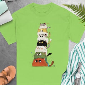 May include: Lime green t-shirt featuring a stack of cartoon cats in various colors. The cats are arranged one on top of the other, creating a whimsical design. The shirt is made of a soft material and is suitable for casual wear.