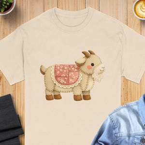 May include: A cream-coloured t-shirt with a whimsical illustration of a goat. The goat has a beige body, brown hooves, and a pink floral saddle. The design is stitched with a decorative border.