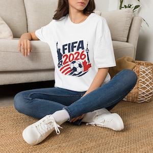 May include: A white short-sleeved t-shirt featuring a graphic print with the text FIFA 2026, a soccer ball, the Statue of Liberty, a maple leaf, and the CN Tower in red, white, and dark blue, paired with dark blue denim jeans and white sneakers.