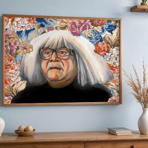 May include: A framed portrait of a person with white hair and glasses, set against a floral background. The artwork features a wooden frame and is displayed on a light blue wall. The portrait is a colorful piece of art.