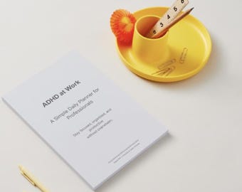May include: A white planner titled "ADHD at Work" with text. A yellow desk organizer holds a pencil, ruler, and paper clips. An orange decorative element sits on the organizer. Two pens are visible.