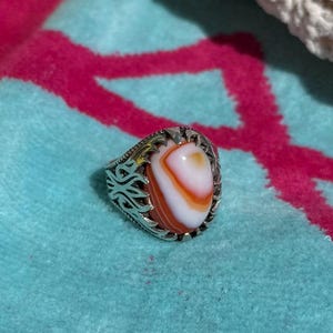 May include: A silver ring with an oval, orange and white stone. The ring has an ornate, filigree design around the stone. The ring is on a blue towel with a pink design, with a white crocheted item in the background.