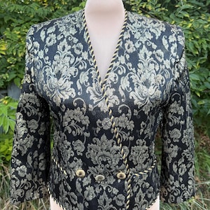 May include: A black blazer featuring a metallic gold floral damask pattern, a V-neckline, and three-quarter length sleeves. The garment is accented with gold braided trim along the edges and includes three round gold buttons at the waist.