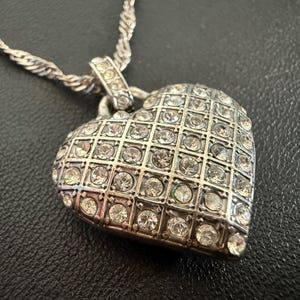 Puede incluir: Collar con colgante de coraz&oacute;n plateado. El colgante en forma de coraz&oacute;n est&aacute; incrustado con numerosos cristales transparentes, dispuestos en un patr&oacute;n de rejilla. La cadena tiene un dise&ntilde;o de cuerda trenzada. El collar se muestra sobre una superficie oscura.