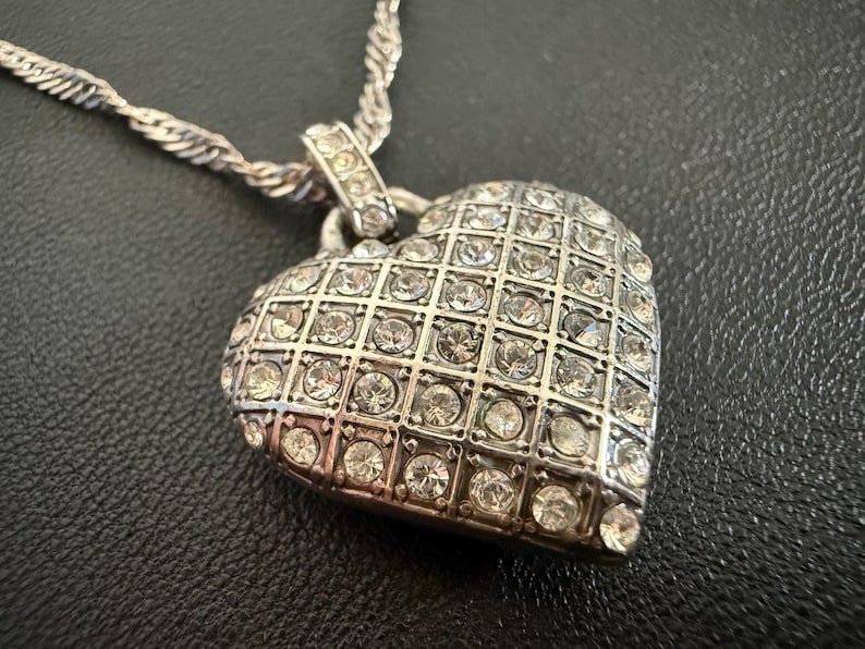 Puede incluir: Collar con colgante de coraz&oacute;n plateado. El colgante en forma de coraz&oacute;n est&aacute; incrustado con numerosos cristales transparentes, dispuestos en un patr&oacute;n de rejilla. La cadena tiene un dise&ntilde;o de cuerda trenzada. El collar se muestra sobre una superficie oscura.