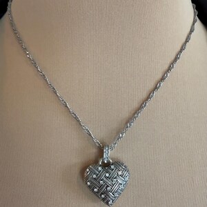 Puede incluir: Collar plateado con colgante en forma de coraz&oacute;n. El colgante presenta un dise&ntilde;o tejido con peque&ntilde;as piedras preciosas transparentes. La cadena es de estilo cuerda trenzada. El collar se muestra sobre un fondo neutro.