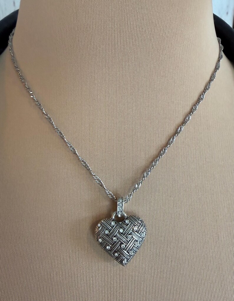 Puede incluir: Collar plateado con colgante en forma de coraz&oacute;n. El colgante presenta un dise&ntilde;o tejido con peque&ntilde;as piedras preciosas transparentes. La cadena es de estilo cuerda trenzada. El collar se muestra sobre un fondo neutro.