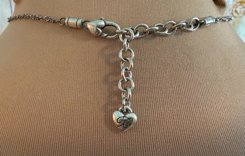 Puede incluir: Primer plano del cierre y la cadena de un collar plateado. El collar presenta un cierre de mosquet&oacute;n, una serie de peque&ntilde;os eslabones de cadena y un dije en forma de coraz&oacute;n con la letra estilizada "B". El collar se muestra sobre una superficie beige.
