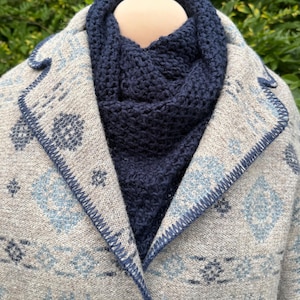 May include: A beige coat featuring a light blue and dark blue geometric pattern with a dark blue blanket stitch along the lapels. A dark navy blue knitted cowl neck scarf is worn underneath the coat, which is fastened with a single silver button.