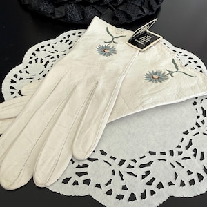 May include: A pair of off-white leather gloves with embroidered blue and green floral accents. The gloves are displayed on a white paper doily with a scalloped edge. A tag is attached to the gloves.