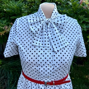 May include: A white short-sleeved dress featuring a black polka dot pattern, a high collar with a large tied bow at the neck, a front button-down closure, and a thin red belt with a silver-toned buckle cinched around the waist.