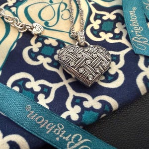 Puede incluir: Un collar con colgante de coraz&oacute;n plateado con un dise&ntilde;o texturizado y piedras transparentes incrustadas. El collar descansa sobre una tela azul y blanca estampada, con una cinta turquesa que dice "Brighton".