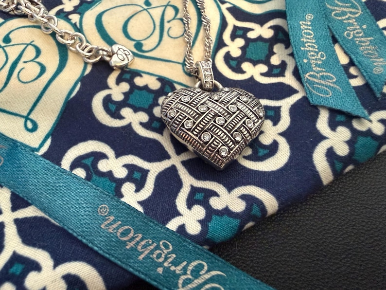 Puede incluir: Un collar con colgante de coraz&oacute;n plateado con un dise&ntilde;o texturizado y piedras transparentes incrustadas. El collar descansa sobre una tela azul y blanca estampada, con una cinta turquesa que dice "Brighton".