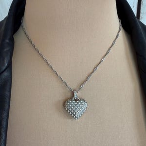 Puede incluir: Collar plateado con colgante en forma de coraz&oacute;n. El colgante est&aacute; incrustado con peque&ntilde;os cristales transparentes dispuestos en un patr&oacute;n de rejilla. La cadena tiene un dise&ntilde;o de cuerda trenzada. El collar se muestra en un maniqu&iacute;.