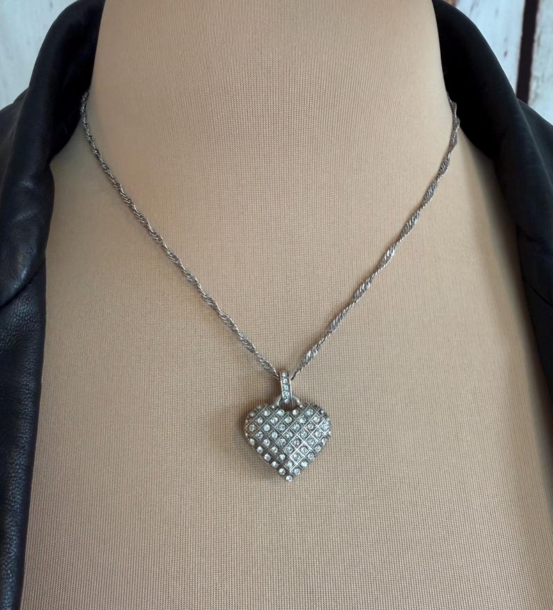 Puede incluir: Collar plateado con colgante en forma de coraz&oacute;n. El colgante est&aacute; incrustado con peque&ntilde;os cristales transparentes dispuestos en un patr&oacute;n de rejilla. La cadena tiene un dise&ntilde;o de cuerda trenzada. El collar se muestra en un maniqu&iacute;.