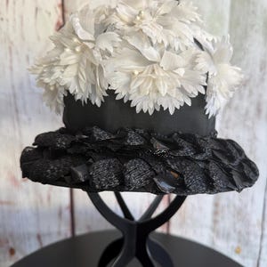 May include: A vintage black hat with a wide brim and a decorative top. The hat features a black ribbon band and is adorned with white fabric flowers. The hat is displayed on a black stand.