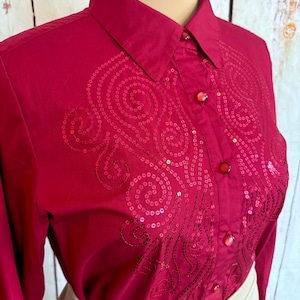 May include: A close-up of a red button-down shirt with long sleeves. The shirt features a sequined design on the front and red buttons. The collar is pointed, and the shirt is partially unbuttoned.
