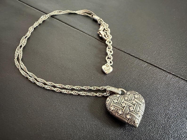 Puede incluir: Collar con colgante de coraz&oacute;n plateado con un dise&ntilde;o tejido y peque&ntilde;as piedras transparentes. El collar tiene una cadena delicada y un peque&ntilde;o dije de coraz&oacute;n cerca del cierre. El colgante mide aproximadamente 2,5 cm de ancho.