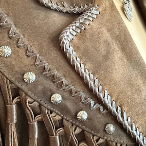 May include: Close-up of a brown suede jacket with decorative silver studs and fringe detailing. The jacket features braided trim along the edges and a matching brown leather necklace with a silver pendant.