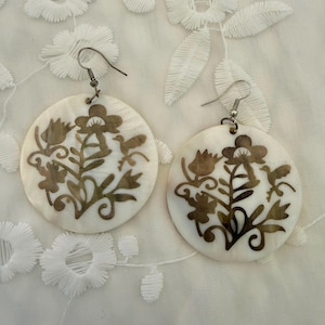 May include: A pair of round, white mother-of-pearl earrings featuring a dark brown, intricate cutout design of flowers, leaves, and a small bird. Each earring hangs from a silver-toned metal hook, showcasing a flat, circular shape with detailed engravings.