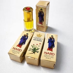 May include: An assortment of NARD perfume bottles and boxes. The bottles are gold with red tops, containing a yellow liquid. The boxes feature an illustration of a woman in blue and red robes, with the text "NARD" and "100% Pure From the HOLY LAND".