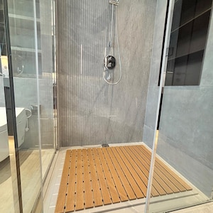 Puede incluir: Una moderna cabina de ducha con una estera de suelo de madera. La ducha cuenta con un cabezal de ducha y accesorios plateados contra una pared de azulejos grises. La cabina está cerrada por paneles de vidrio transparente.