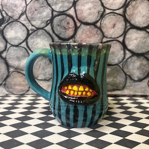 May include: A ceramic mug with a teal handle and vertical teal and black stripes. The mug features a black mouth with yellow teeth. The mug is on a black and white checkered surface.