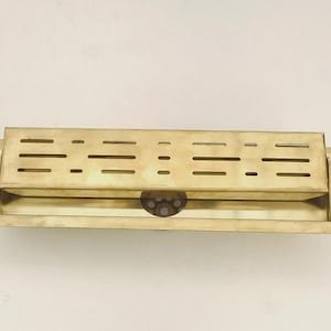 May include: A rectangular brass object with a series of horizontal slots and a hinged lid. The brass has a polished finish, reflecting light. The object appears to be a decorative or functional item, possibly for holding small items.