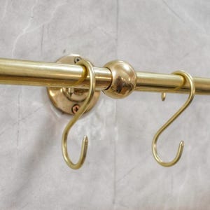 May include: A brass kitchen rail with S-shaped hooks. The rail is mounted on a wall and has a decorative ball in the centre. The hooks are designed for hanging kitchen utensils or other items.