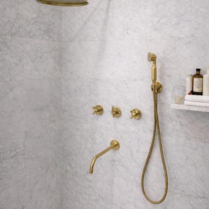 Puede incluir: Juego de grifería de ducha dorada contra una pared de azulejos de mármol. El juego incluye un cabezal de ducha de lluvia, una ducha de mano y controles de grifo. Los accesorios tienen un diseño clásico.