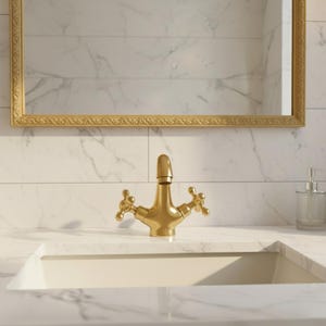May include: A gold-colored bathroom faucet with cross-shaped handles is centered above a white sink. A gold-framed mirror hangs above the faucet. A clear soap dispenser sits to the right of the sink. The background is white marble tile.