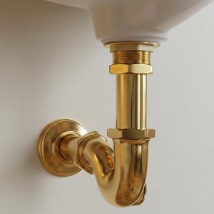 May include: A close-up of a brass-colored plumbing fixture, including a P-trap and connecting pipes, installed beneath a white sink. The fixture has a polished finish and is mounted against a neutral-toned wall.