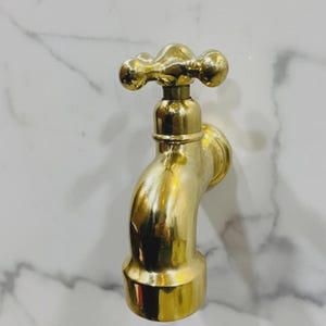 May include: A polished gold-coloured tap with a cross-shaped handle. The tap has a curved spout and is mounted on a white marble-effect wall. The fixture is made of metal and has a reflective surface.