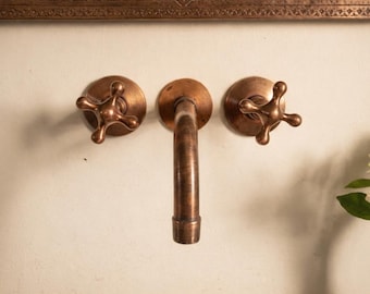 May include: Close-up of a copper-toned bathroom faucet set against a cream-colored wall. The set includes a curved spout and two cross-shaped handles. A white sink with the word "brasska" is visible below.