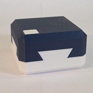 May include: A rectangular box with a dark blue lid and a white base. The lid features a textured surface with a small white square inset on the top corner. The box has a geometric design with angled edges and a distinct interlocking seam pattern.