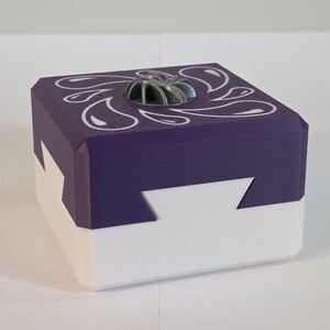 May include: A square box with a white base and a purple lid featuring a white teardrop pattern. The lid has a central circular silver-colored vent with radial slats. The box sides have a geometric interlocking design where the purple lid meets the white base.