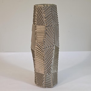 May include: A tall, cylindrical vase featuring a textured surface with a repeating geometric pattern of raised, diagonal lines in a muted taupe and grey color palette. The design consists of distinct panels with alternating line directions.