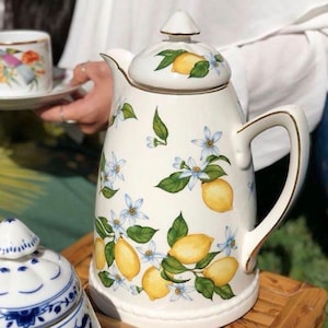 Pode incluir: Uma chaleira de cerâmica branca com tampa, alça e bico. A chaleira é decorada com limões amarelos, folhas verdes e pequenas flores azuis. A borda e a alça têm acabamento dourado. Uma chávena e um pires a condizer estão no fundo.