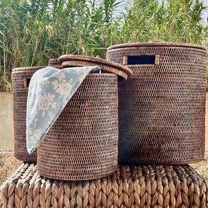 May include: Three brown woven wicker storage baskets of varying sizes. One basket has a lid, and another has a light blue floral patterned cloth draped over the side. The baskets are on a woven surface, with greenery in the background.