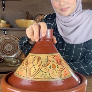 May include: A traditional terracotta tagine pot with a conical lid. The pot is a warm reddish-brown colour, with a decorative yellow and orange pattern on the lid. The base is round and wide.