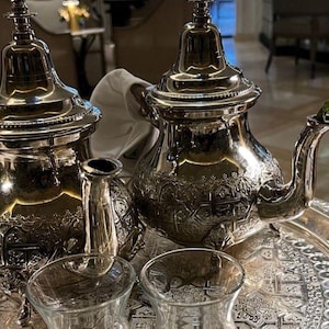Peut inclure: Un service à thé argenté avec des détails ornés, comprenant deux théières, un petit pichet et deux verres transparents, le tout disposé sur un plateau argenté décoratif. Les théières présentent des motifs complexes et des couvercles hauts et pointus.