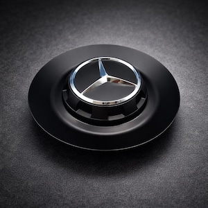 May include: A black hubcap featuring the Mercedes-Benz logo. The logo is a silver three-pointed star inside a silver ring, set against a black background. The hubcap has a matte finish and a circular design.
