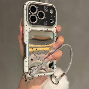 May include: White and silver phone case with a star pattern and a mirrored cardholder. The case features a silver strap with glitter and a white fur pom-pom. The case has a unicorn design and the text "WITH A LITTLE SPARKLE".