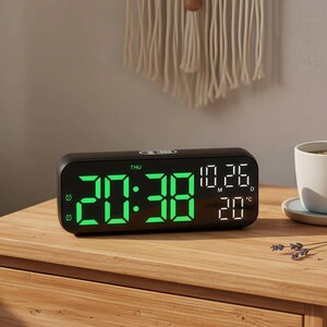 Op de afbeelding: Een zwarte digitale wekker toont tijd, datum en temperatuur in groene LED-verlichting. De wekker staat op een houten nachtkastje naast een witte theekop en schotel. Een decoratieve wandophanging is op de achtergrond.