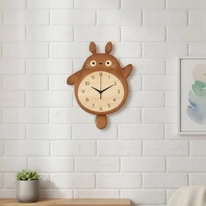 May include: A brown wooden wall clock shaped like a cartoon character, with a light wood clock face and black hands. The clock is mounted on a white brick wall, with a shelf and artwork below.